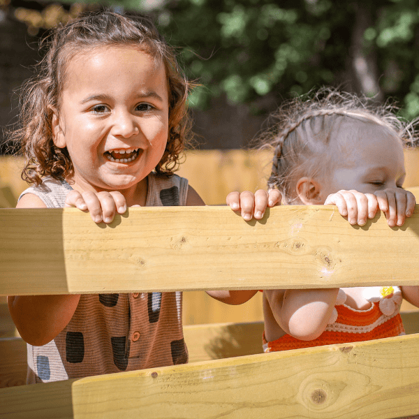 Toddlers Playing Outdoors at Monkey Puzzle Two happy toddlers playing outdoors behind a wooden fence at Monkey Puzzle Day Nursery.